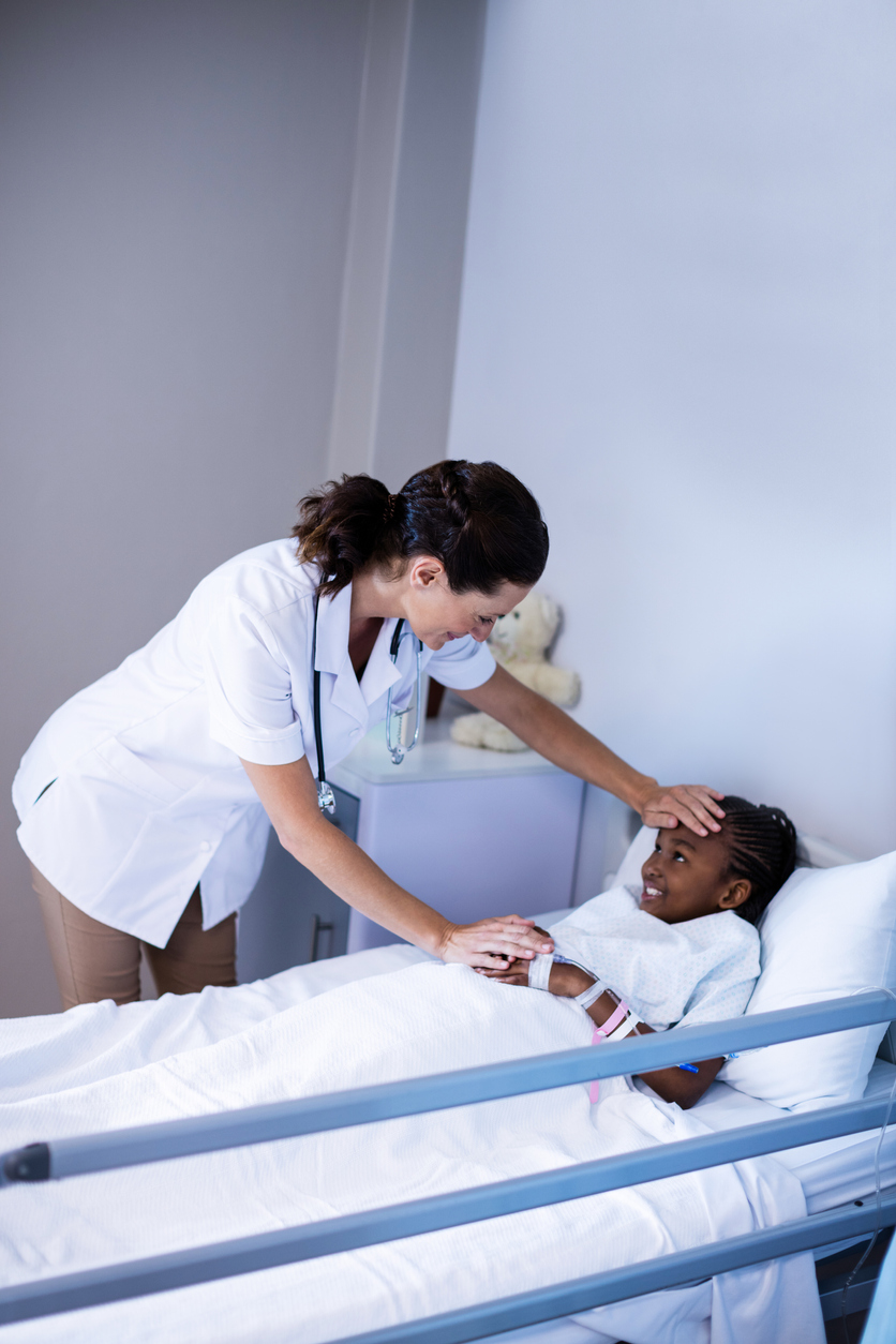 Black child in hospital bed with nurse with long curly black hair in a ponytail leaned over the bed