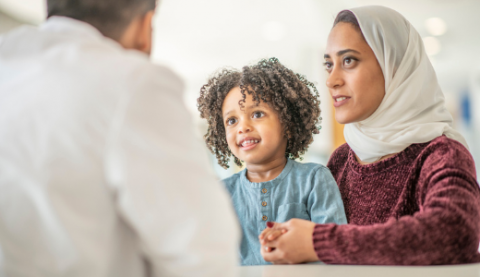 Woman wearing white hijab and maroon top with child with curly hair, both dark brown skin talking to physician with short salt and pepper gray and black hair