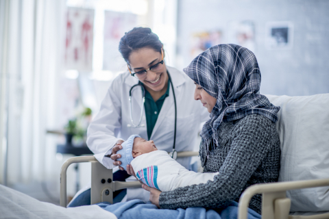 Doctor with long dark hair pulled back into ponytail, glasses, and stethoscope around her neck talks to person in hijab with baby  