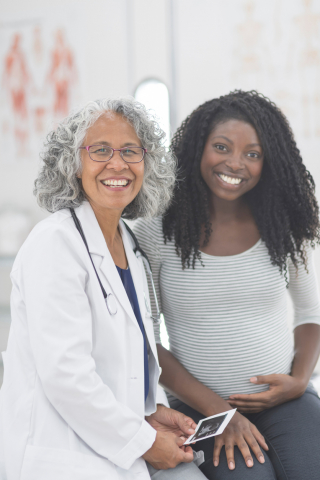 Black woman doctor with gray kinky curly hair wearing lab coat and glasses, smiles next to pregnant dark-skinned Black woman with black long curly hair wearing gray shirt